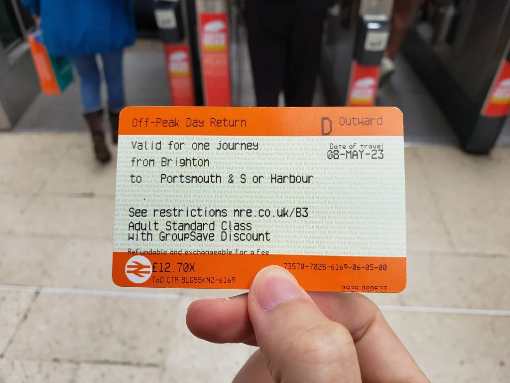 Un billet de train orange National Rail tenu devant les portiques d'une gare